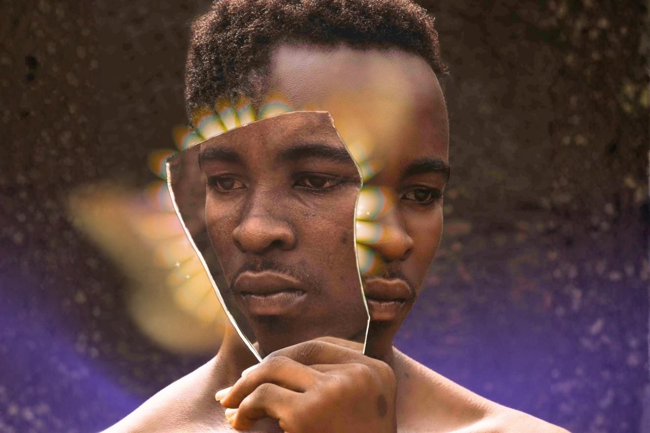 Self-Consent. The image shows a man holding a broken mirror shard that reflects his own face, creating a fragmented and introspective visual effect. The background is dark and textured with soft golden and purple light flares radiating from behind the mirror, giving the composition a surreal, dreamlike quality. The warm brown skin tones contrast against the cool, ethereal lighting, emphasizing themes of reflection, identity, and self-perception.