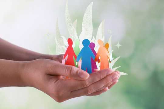 Sex differences. The image features a pair of hands gently cradling a group of colorful paper-cutout figures representing people. The figures are in various colors, including red, blue, green, pink, orange, and purple, symbolizing diversity and unity. Behind the figures, there are subtle, leafy shapes and sparkles, adding a touch of softness and magic to the background. The background itself is a light, blurred green, evoking a sense of nature and harmony. The composition emphasizes themes of care, community, and inclusivity.