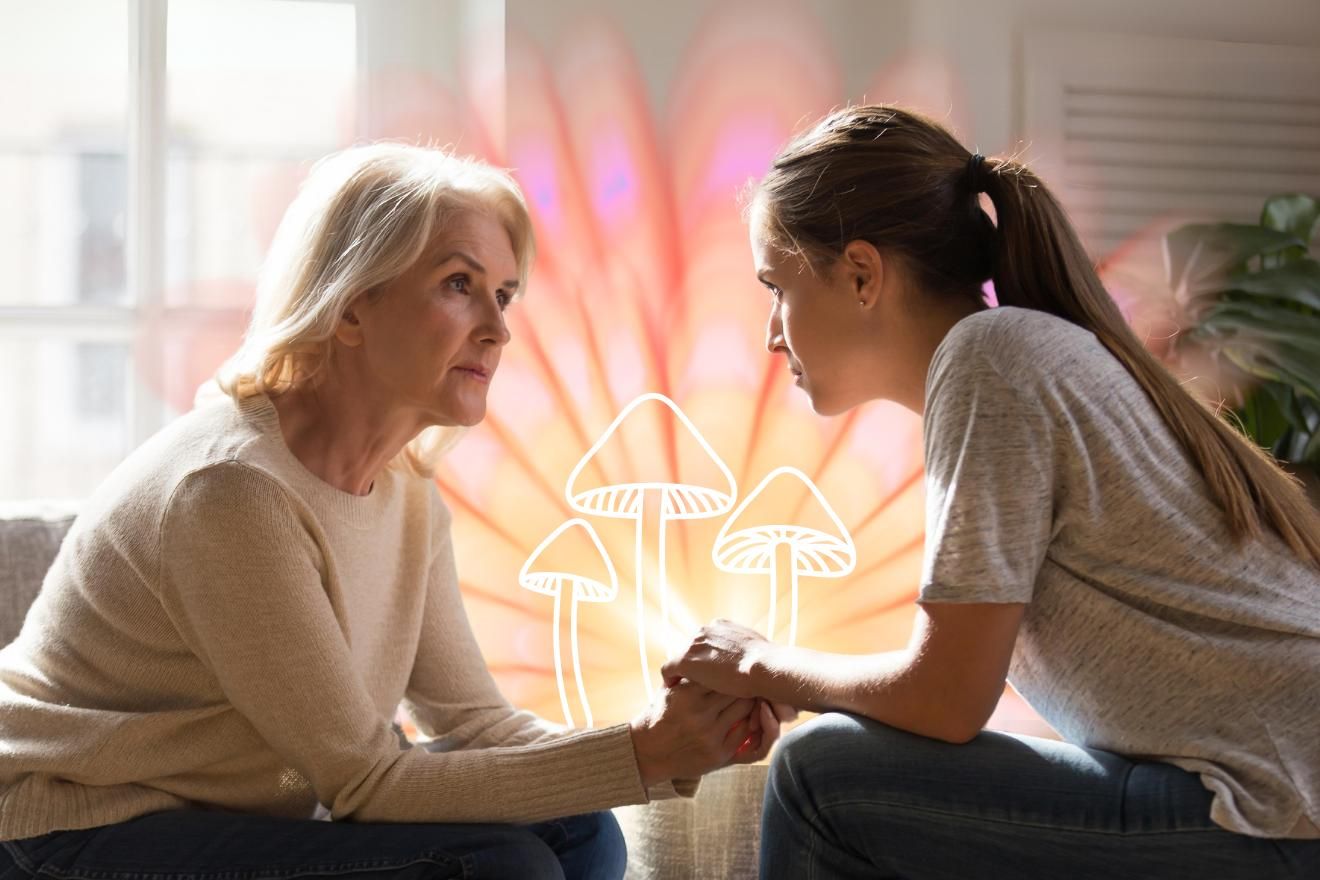 Perimenopause. A photograph of two women sitting across from one another holding hands. The woman on the left is older and wearing a long-sleeved top with light shoulder-length hair. The woman on the right is younger and wearing a t-shirt with dark hair tied up in a ponytail. Behind them is a glowing floral-shaped light in sunset colors, with graphics of psilocybin mushrooms in thin white outlines in the background space between the two women.