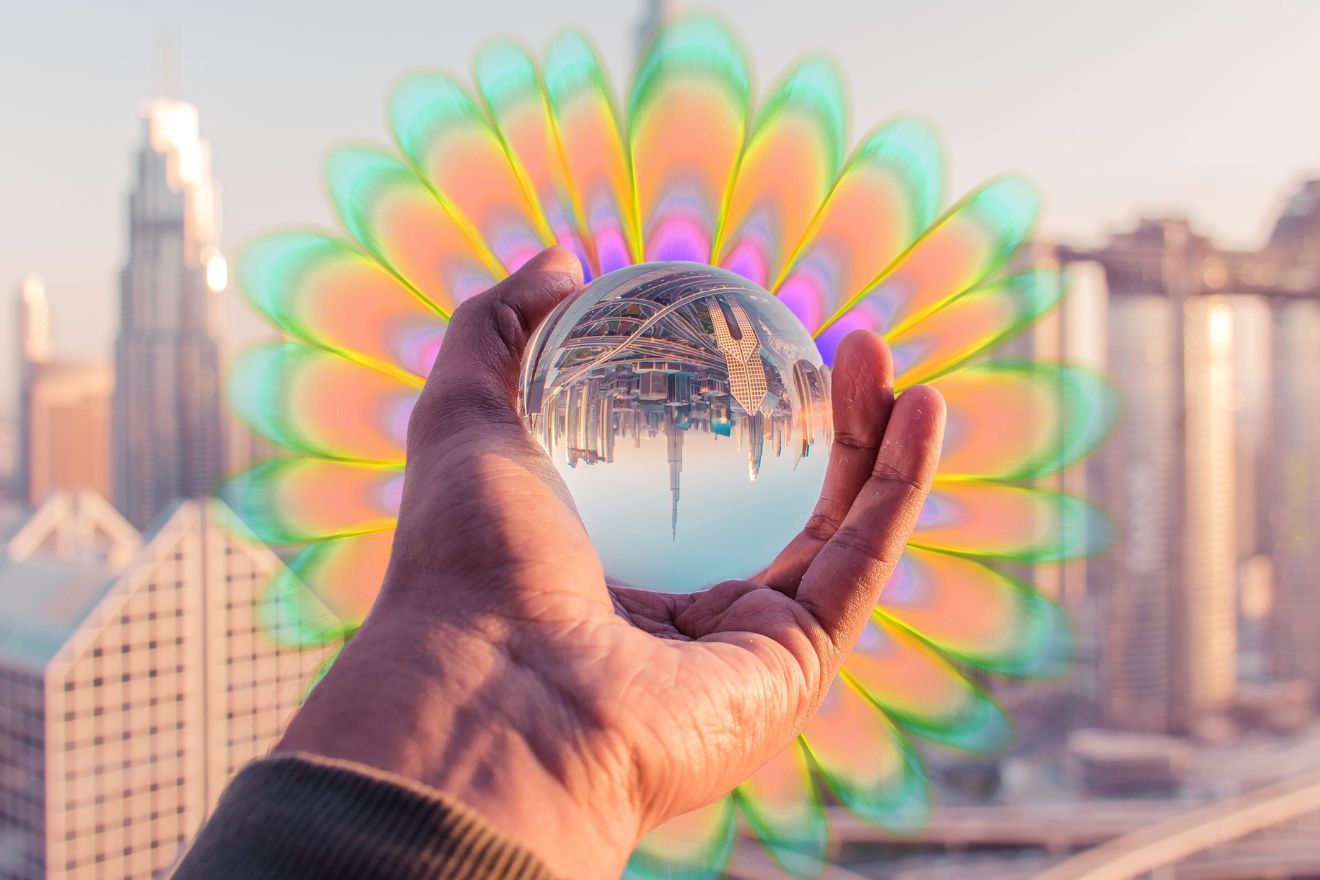 Psychedelic Business. Image is of an out-of-focus cityscape, with a person's hand in the foreground. The hand is holding an orb, that has the same cityscape in focus, on a smaller scale within the orb, and upside down. Behind the person's hand is a flower-shaped pattern with layers in different colors, appearing as if to emanate from the orb.